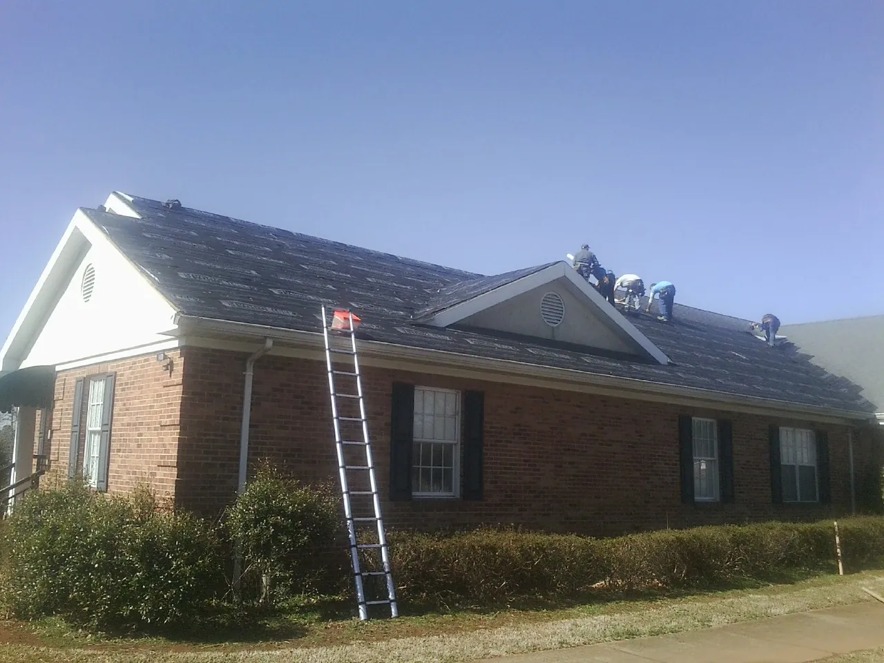 Crew tearing shingles off a brick ranch roof with a ladder set — Metal Roof Repair work by our crew in Maryknoll