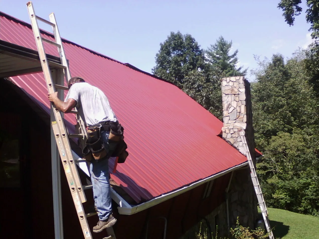 Roofer on a ladder beside a red metal roof and stone chimney — Soffit & Fascia Repair by Roofing Brothers in Maryknoll